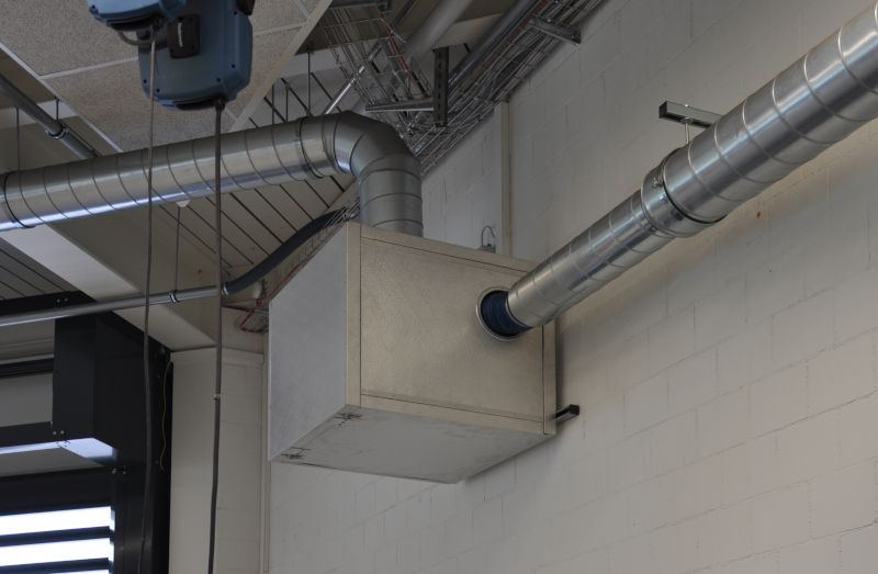 Wandmontierter rechteckiger Filterkasten, verbunden mit runden Metallrohren einer Lüftungs- oder Absauganlage.