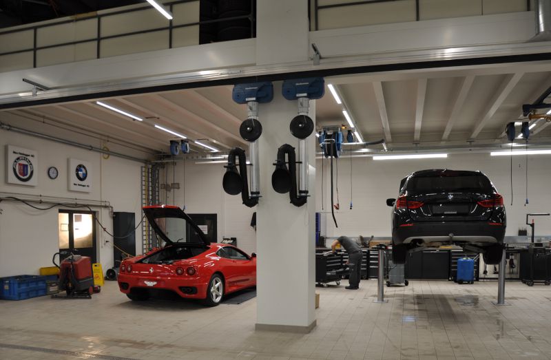 Werkstatt mit zwei Fahrzeugen, eines auf einer Hebebühne. Schlauchaufroller an Säule und Decke sichtbar.