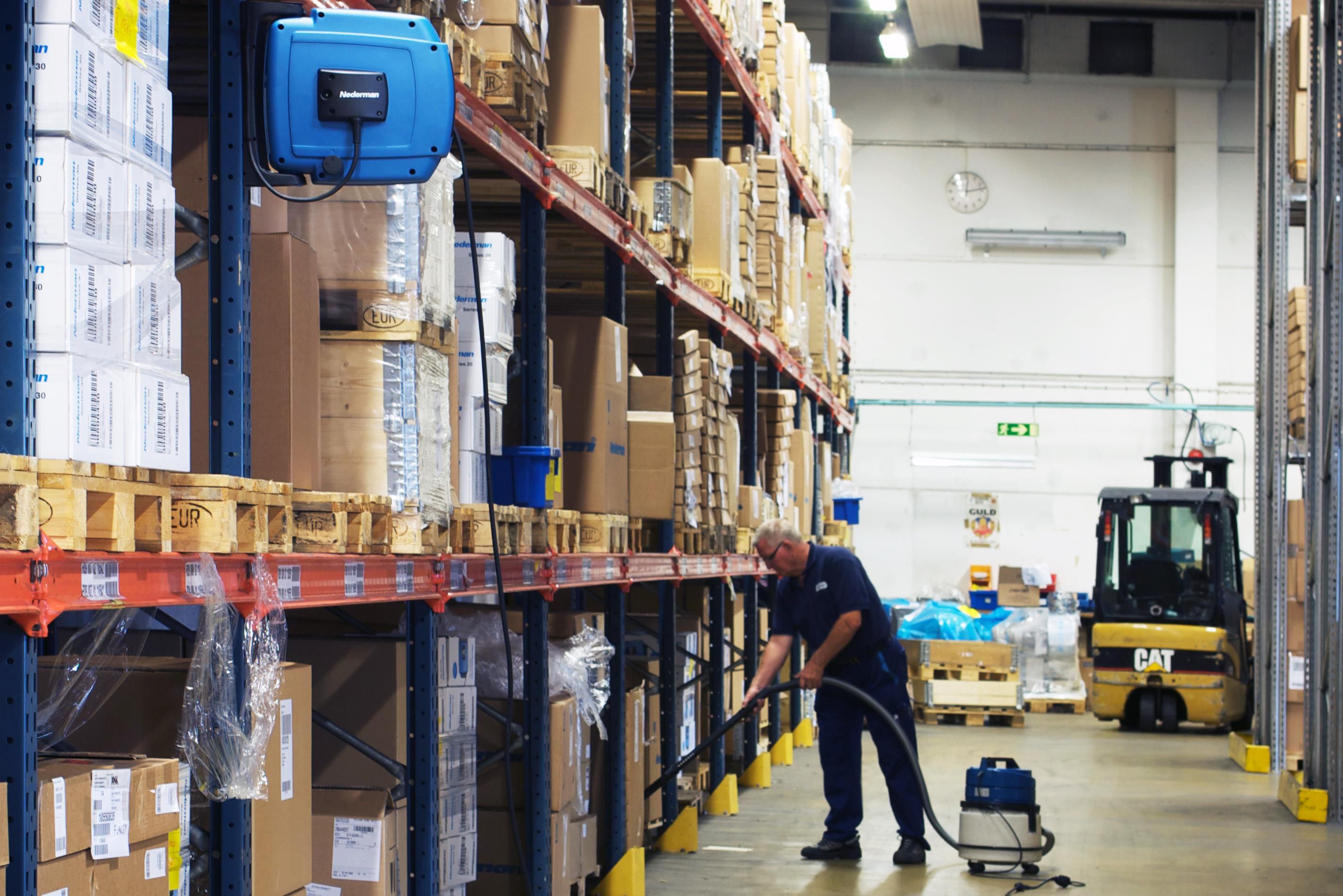 Enrouleur de câble Nederman C30 monté sur un support dans un entrepôt ; en dessous, un homme équipé d'un aspirateur connecté procède au nettoyage.